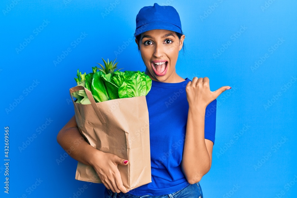 Beautiful brunette woman wearing courier uniform with groceries bag pointing thumb up to the side smiling happy with open mouth Beautiful brunette woman wearing courier uniform with groceries bag pointing thumb up to the side smiling happy with open mouth