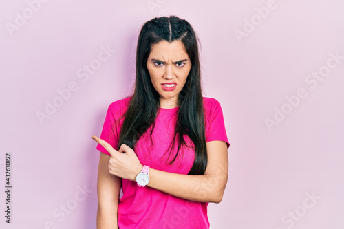 Young hispanic girl wearing casual pink t shirt pointing aside worried and nervous with forefinger, concerned and surprised expression