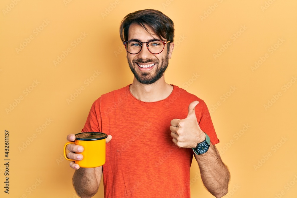 Young hispanic man drinking a cup of coffee smiling happy and positive, thumb up doing excellent and approval sign Young hispanic man drinking a cup of coffee smiling happy and positive, thumb up doing excellent and approval sign
