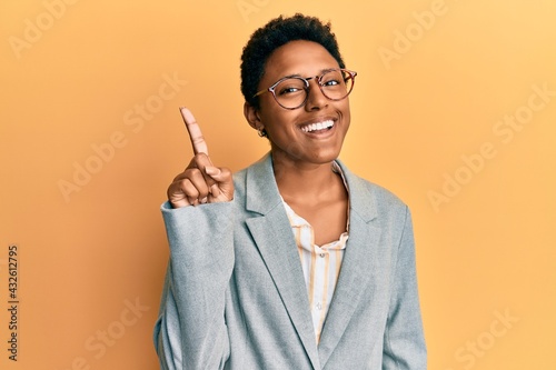 Young african american girl wearing business jacket and glasses showing and pointing up with finger number one while smiling confident and happy. photo