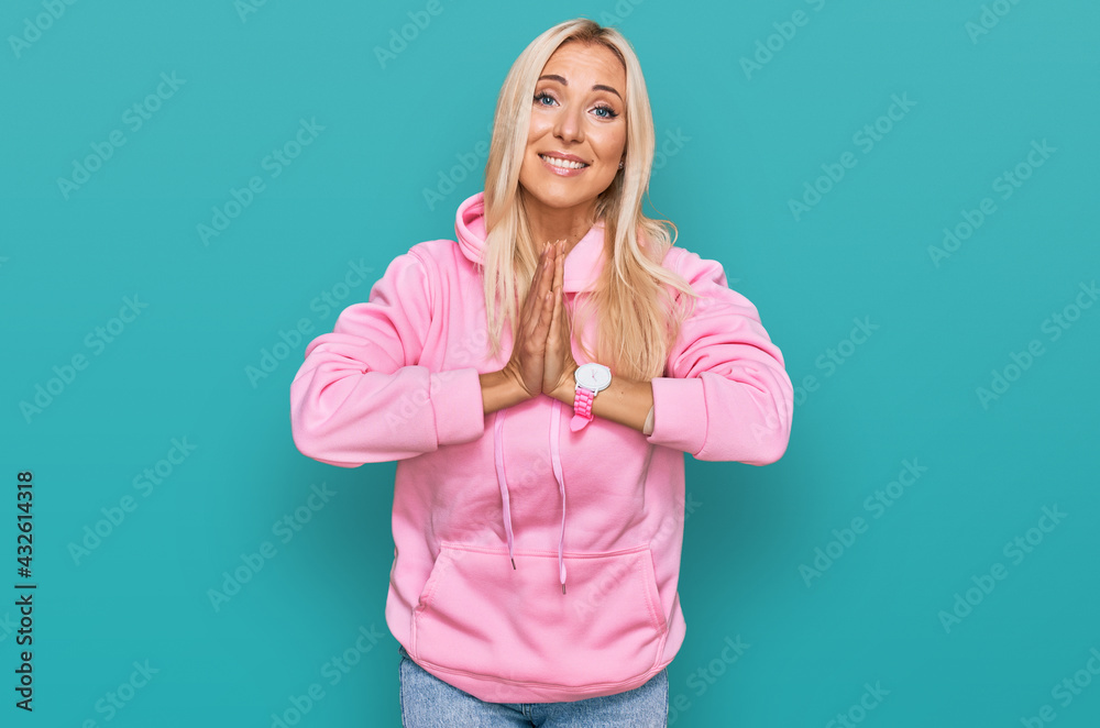 Young blonde woman wearing casual sweatshirt praying with hands together asking for forgiveness smiling confident.