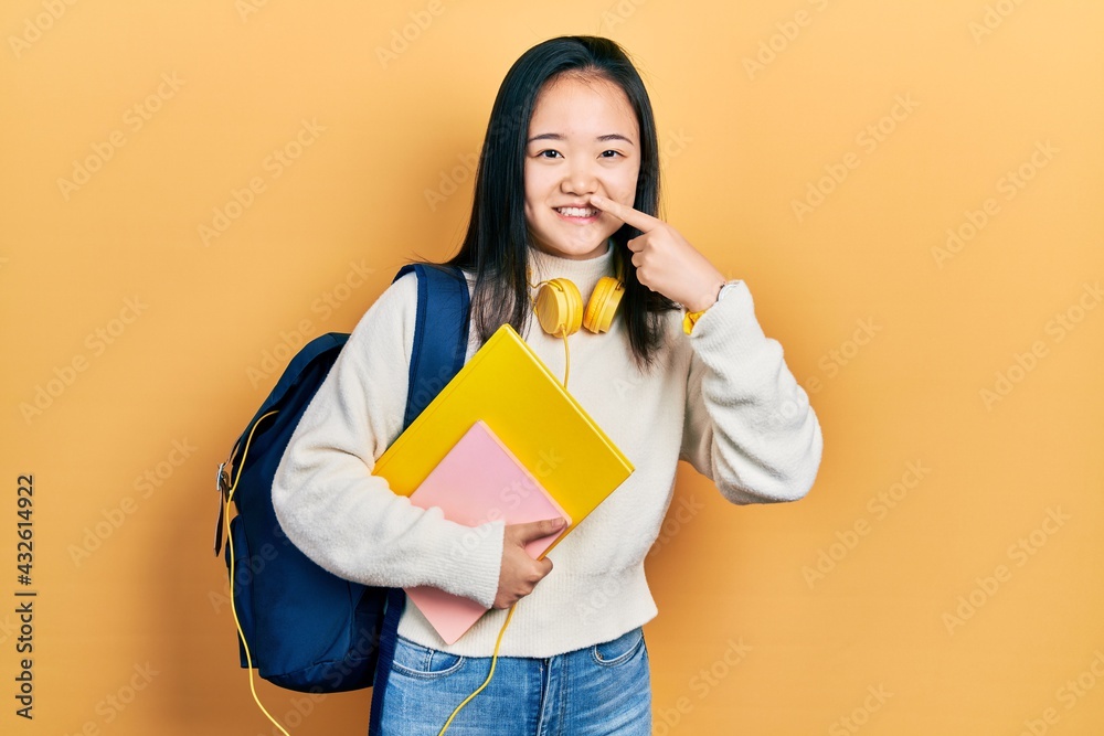 Young chinese girl holding student backpack and books pointing with hand finger to face and nose, smiling cheerful. beauty concept Young chinese girl holding student backpack and books pointing with hand finger to face and nose, smiling cheerful. beauty concept