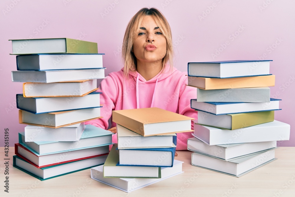 Young caucasian woman sitting on the table with books looking at the camera blowing a kiss on air being lovely and sexy. love expression. Young caucasian woman sitting on the table with books looking at the camera blowing a kiss on air being lovely and sexy. love expression.