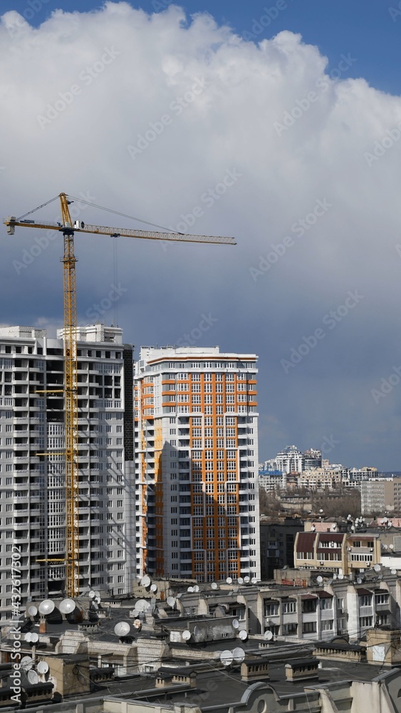 Construction site of unfinished multistory building with crane jib with cumulus cloud in sky. Under construction cityscape with rooftops and cloudscape. Vertical shot