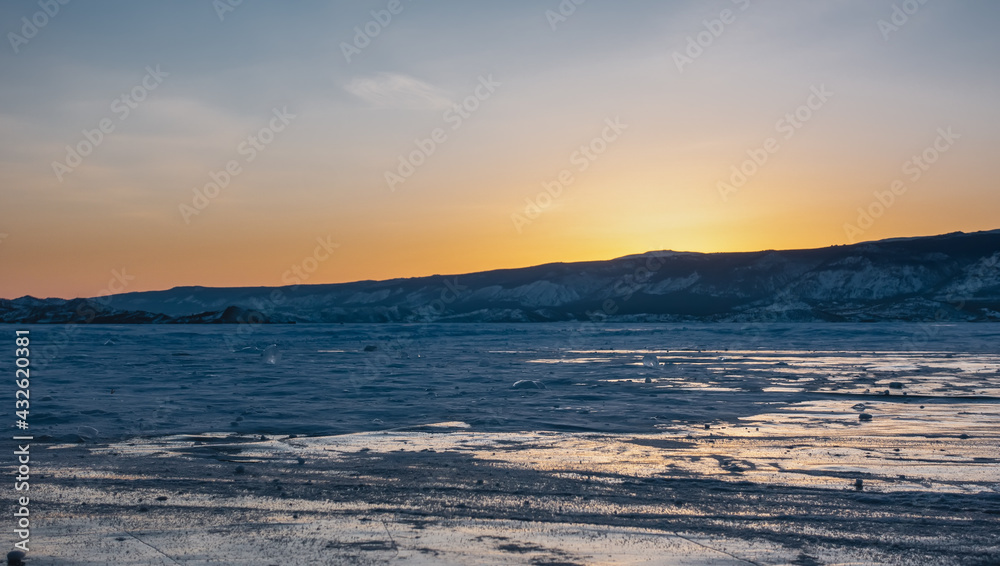 Sunset over a frozen lake. The sky above the mountain range is colored orange. On the ice there are patches of snow and reflections of the setting sun. Baikal