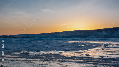 Sunset over a frozen lake. The sky above the mountain range is colored orange. On the ice there are patches of snow and reflections of the setting sun. Baikal © Вера 