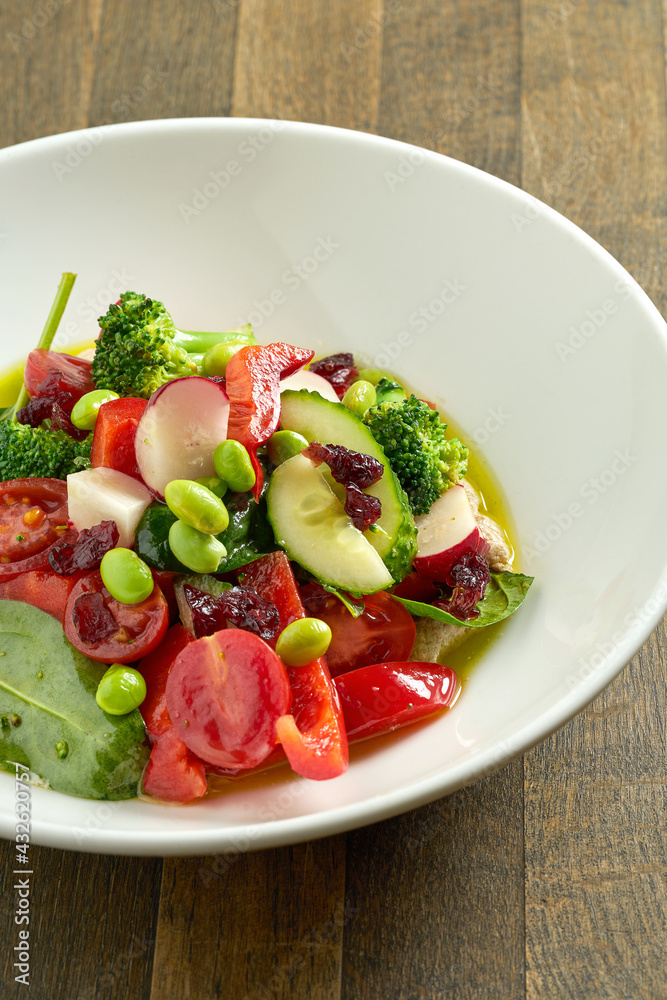Appetizing vegetable salad with cucumber, tomatoes, radish, beans and broccoli with green butter in a white bowl on a wooden background. Delicious, dietary and vegetarian food