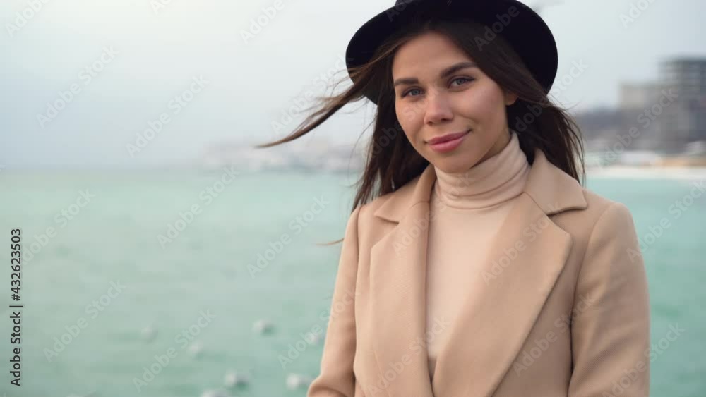 Close-up portrait, a young Caucasian girl stands on the pier dressed in a coat and hat near the ocean or sea and looks into the camera. Copy space. Close-up portrait, a young Caucasian girl stands on the pier dressed in a coat and hat near the ocean or sea and looks into the camera. Copy space.