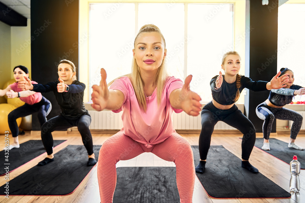 Obraz premium Young woman doing squat exercise at group fitness training with other people girls standing in row wearing sportswear performing crouching at gym. 