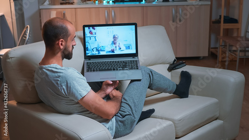 Doctor consulting sick patient during coronavirus quarantine using laptop computer for videocall consultation sitting on sofa. Caucasian man disscusing about illness symptoms and headlcare treatment photo