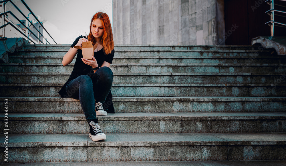 young woman or teenage girl eating asian fast food from takeaway box sitting on the steps on city street. Thai noodles in paper box takeaway street food young woman or teenage girl eating asian fast food from takeaway box sitting on the steps on city street. Thai noodles in paper box takeaway street food