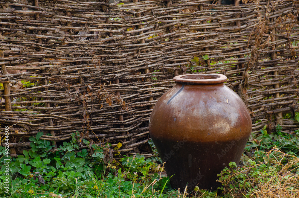 Pot at wicker fence at countryside. Antique traditional tableware. Rural landscapes. Old handmade clay jug. Ceramic brown pitcher. Hand manufactured clay pot handcrafted. Pot at wicker fence at countryside. Antique traditional tableware. Rural landscapes. Old handmade clay jug. Ceramic brown pitcher. Hand manufactured clay pot handcrafted.