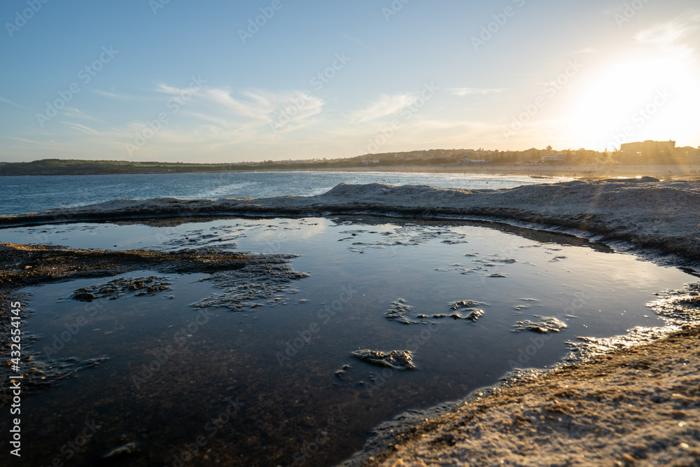 Naklejka premium reflection in a puddle of the sun setting on the rocks at the beach