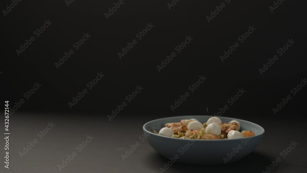 Slow motion ground fresh spices on pesto penne with fried shrimps and mozzarella in blue bowl on black background Slow motion ground fresh spices on pesto penne with fried shrimps and mozzarella in blue bowl on black background
