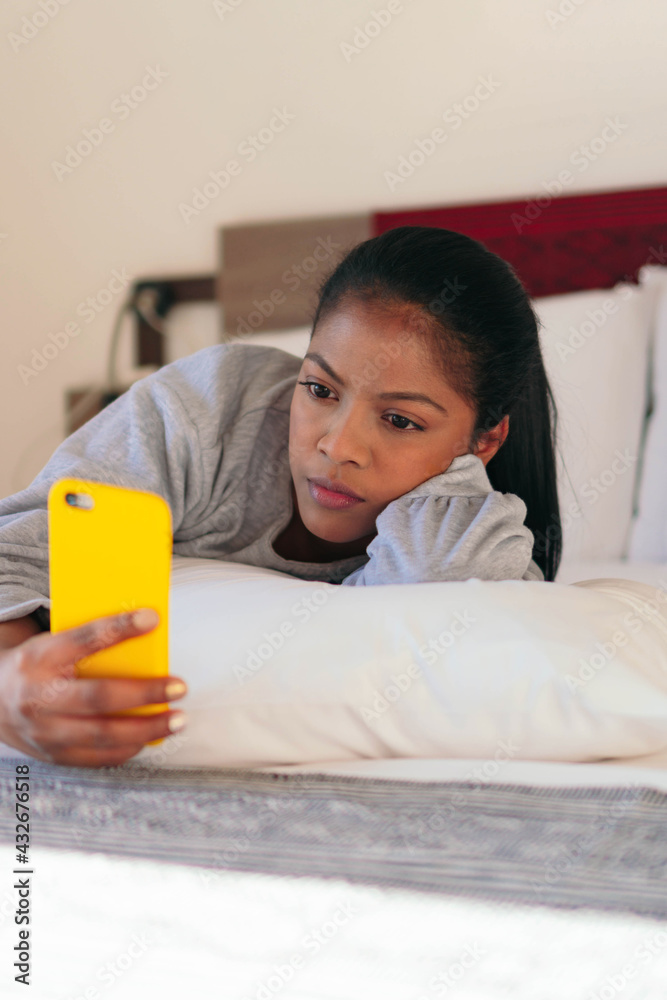 An African American woman holds her phone while lying in bed in her bedroom. An African American woman holds her phone while lying in bed in her bedroom.