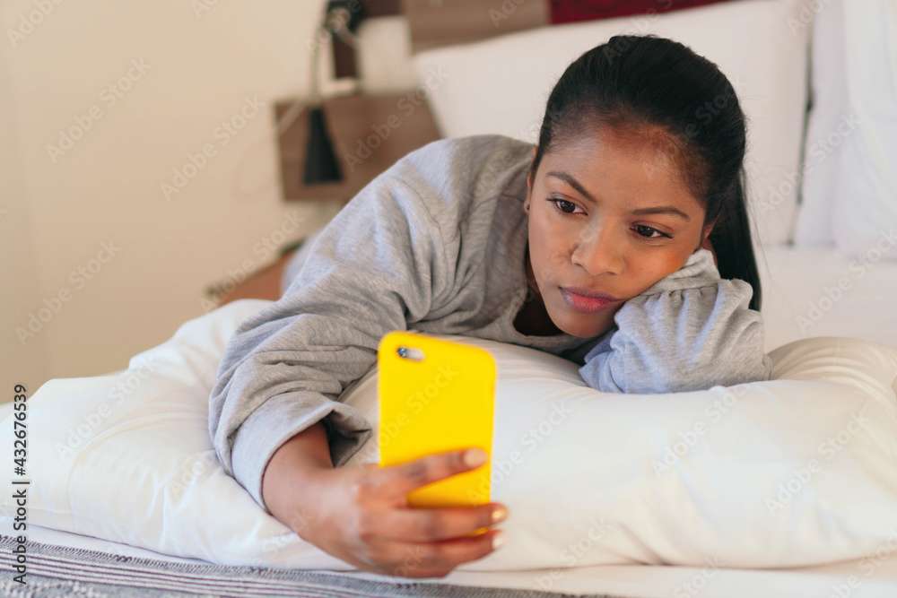 An African woman holds her phone while lying in bed in her bedroom. An African woman holds her phone while lying in bed in her bedroom.