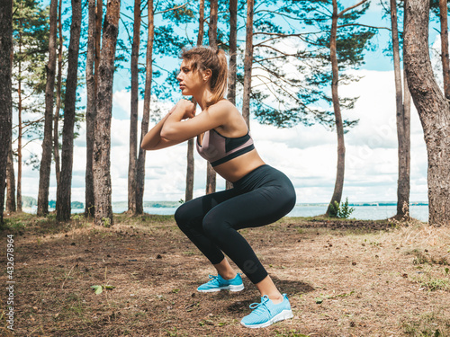 Portrait of concentrated young sports woman in the park. Model makes sport stretching exercises before training outdoors. Female posing in the forest in sportswear.Making squats