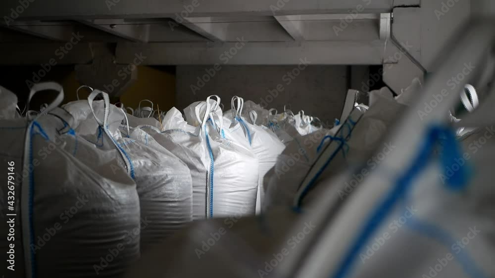 Close-up large, full bags of cereal products in stock. The bags are stacked in rows, industrial warehouse. Big white plastic bags with cereals: rye, wheat, barley, mustard are storing in storage. Close-up large, full bags of cereal products in stock. The bags are stacked in rows, industrial warehouse. Big white plastic bags with cereals: rye, wheat, barley, mustard are storing in storage.