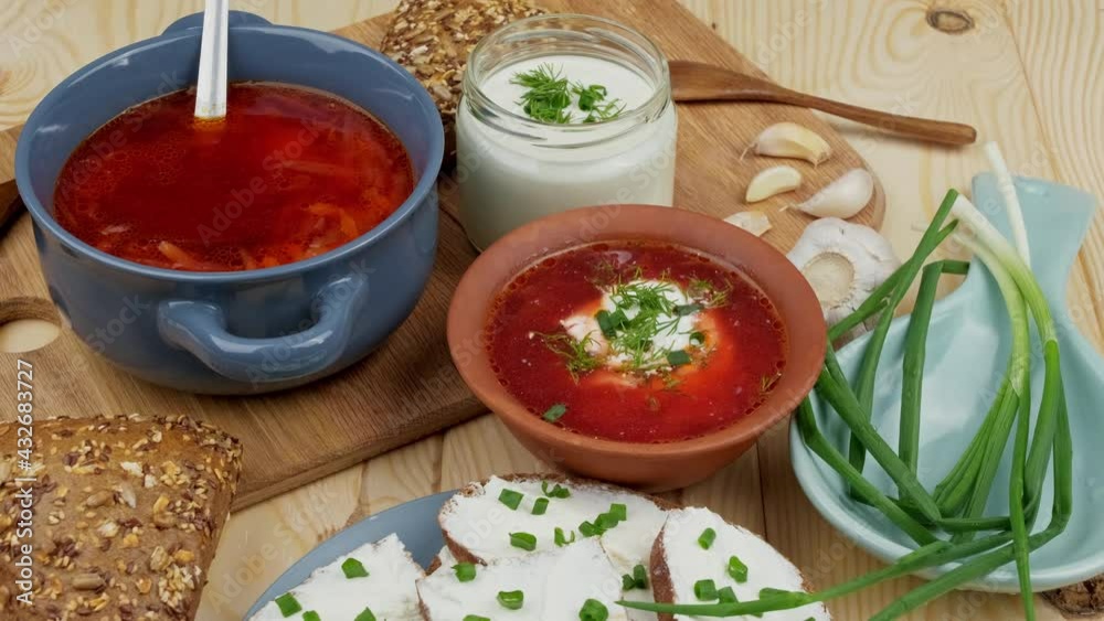 Russian and Ukrainian cuisine. Borscht with sour cream and spices, fresh herbs, garlic and pampushki. Beetroot soup in a blue bowl on a wooden board. Russian and Ukrainian cuisine. Borscht with sour cream and spices, fresh herbs, garlic and pampushki. Beetroot soup in a blue bowl on a wooden board.