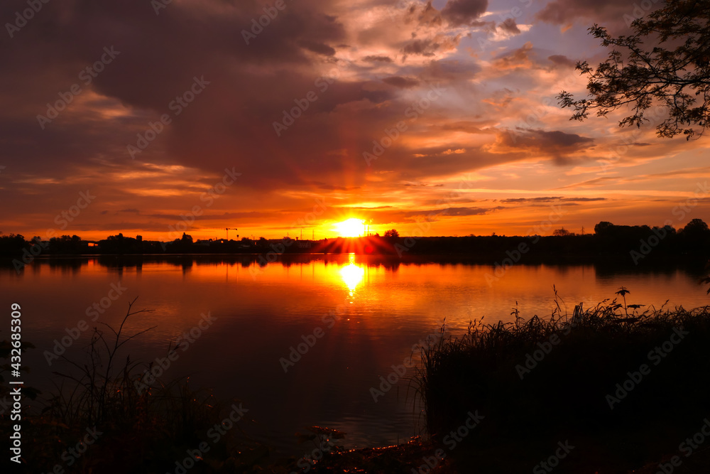 Amazing landscape with dramatic sky over the water. Sunrise or sunset in rural scene with silhouette of vegetation in front of a lake.