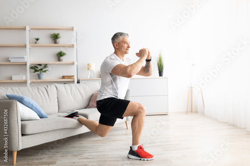 Active mature man doing squats while training at home, doing exercises near sofa in living room interior