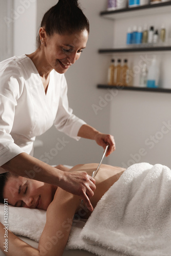 Massage at spa salon. Smiling beautician at beauty clinic using metal gua sha for body back massage, tsm asian procedure to relieve tension and tightness, gouache scrapping