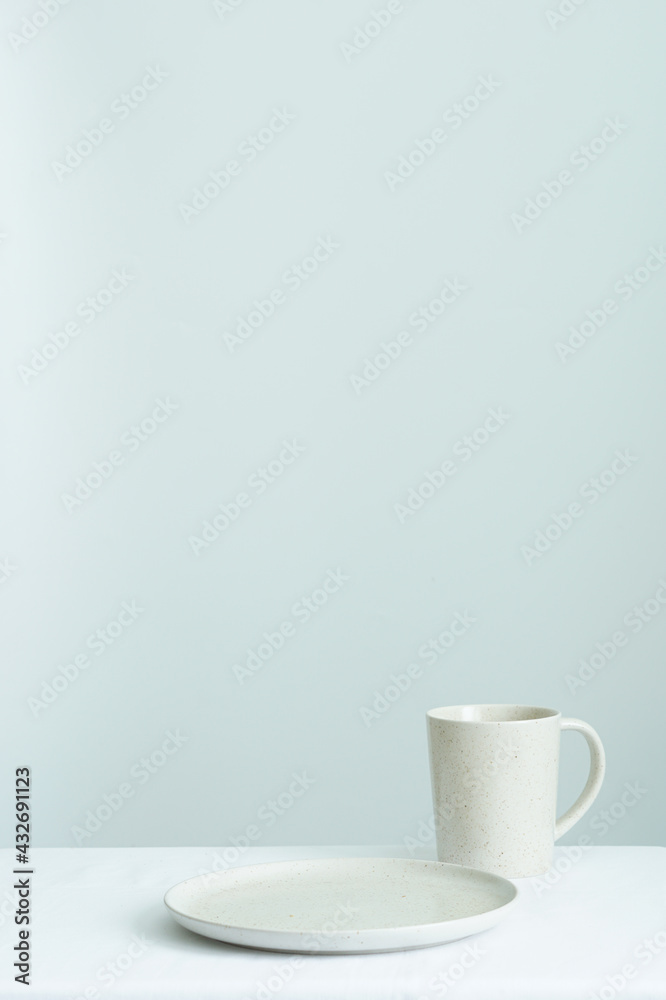 vertical image of empty circle ceramic plate and ceramic mug placed on white tablecloths against white wall