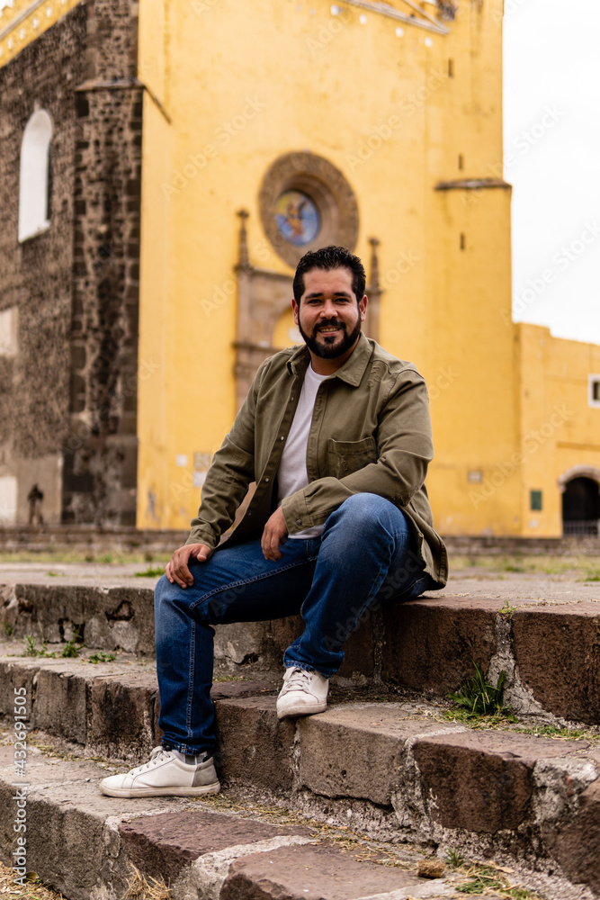 Young mexican bearded guy with a jacket sitting on stone steps in a franciscan convent in Mexico. Full body portrait.