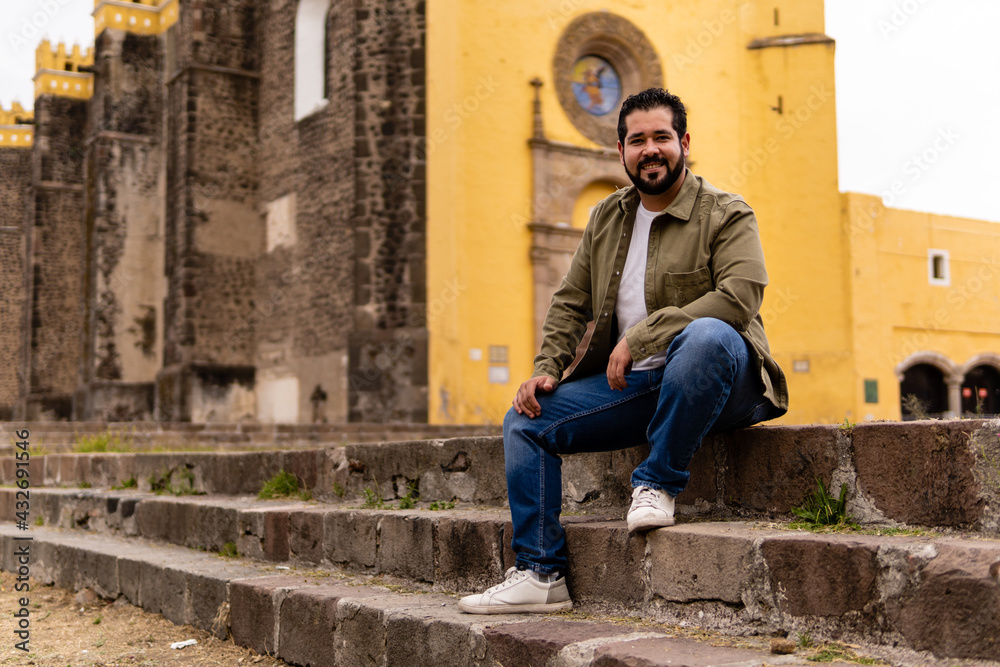 Obraz premium Young mexican bearded guy with a jacket sitting on stone steps in a franciscan convent in Mexico. Full body portrait.