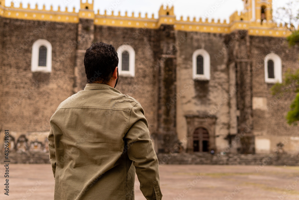 Young bearded mexican man wearing green jacket admiring a church in Mexico. Portrait from the back. 