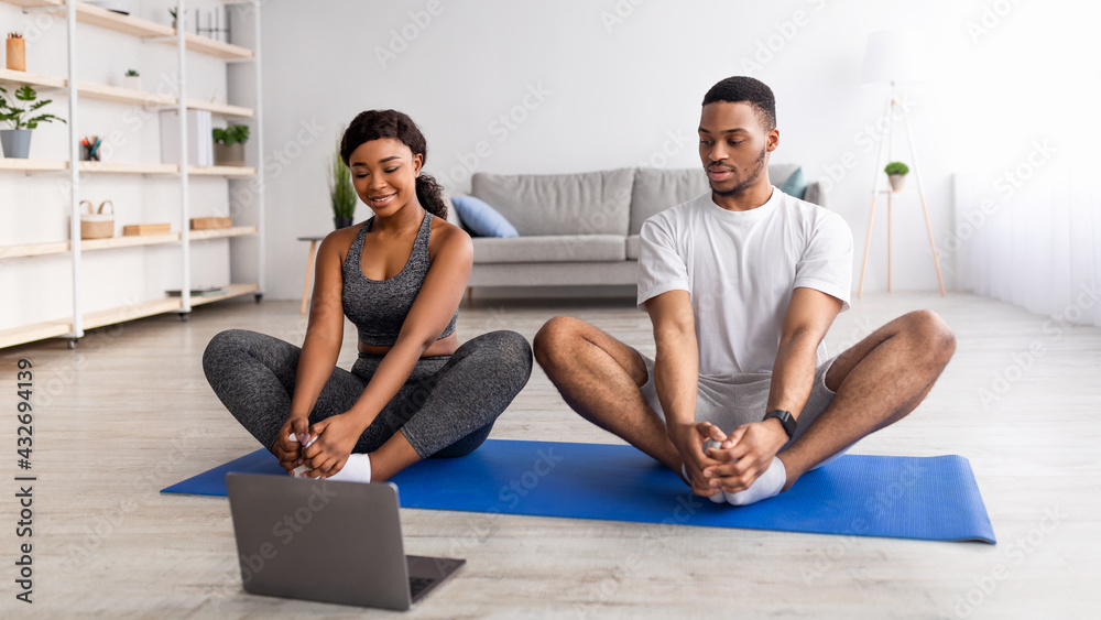 Online yoga practice. Young black couple doing butterfly asana on sports mats, following video tutorial on laptop pc