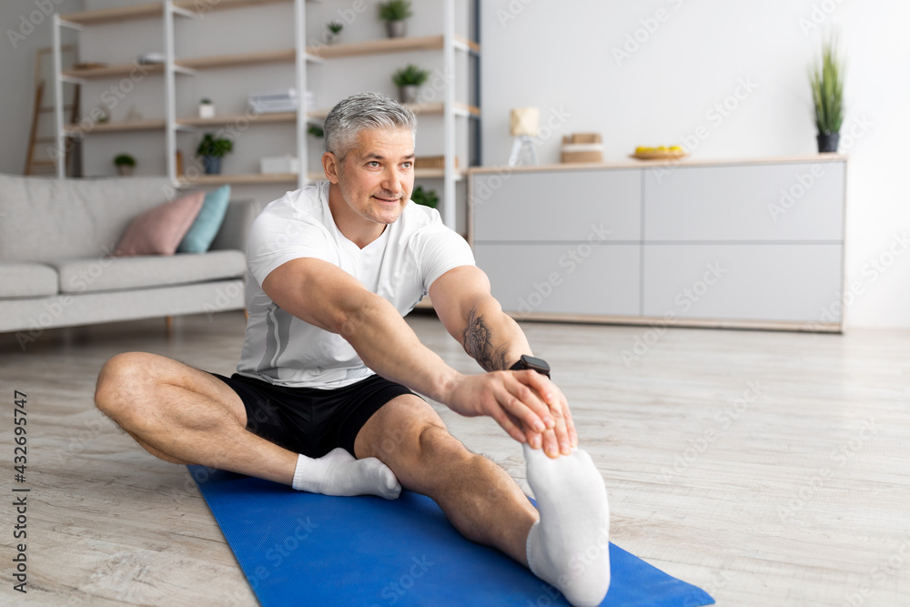 Foto Stock Healthy active lifestyle. Mature man in sportswear and socks,  doing stretching exercises for legs on mat in living room | Adobe Stock