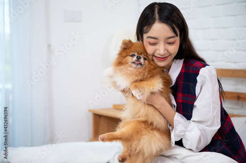 Young pretty Asian woman sitting and embracing Brown Pommeranian dog on white bed with copy space photo