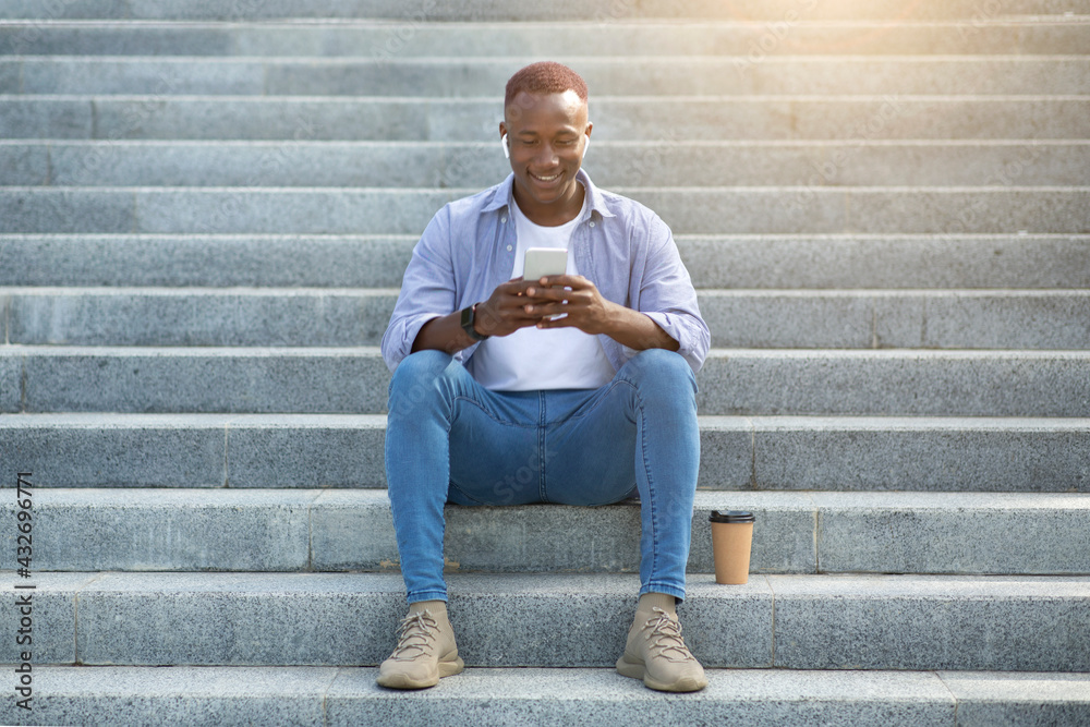 Cheerful African American guy sitting on stairs and browsing internet on mobile phone, outdoors