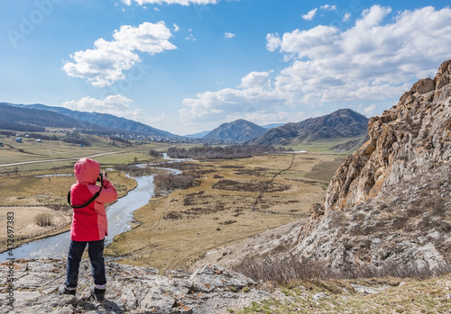 a little girl, a child in an autumn warm jacket stands and takes pictures with a camera of a landscape with a river, standing on a stone of a high mountain