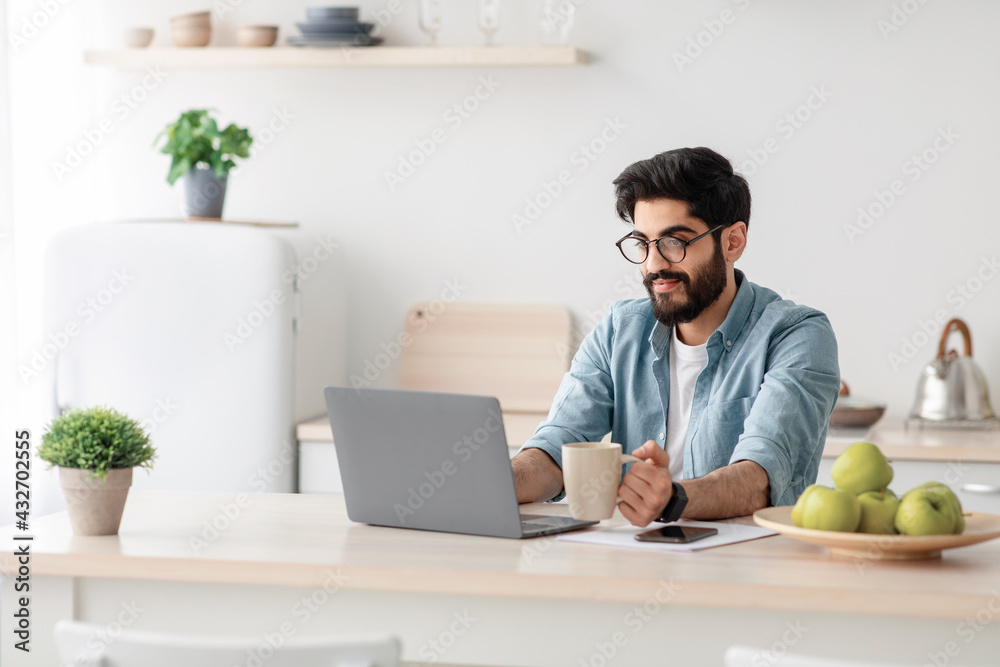 Modern job lifestyle. Happy arab male freelancer working on laptop in kitchen, enjoying distance job opportunities Modern job lifestyle. Happy arab male freelancer working on laptop in kitchen, enjoying distance job opportunities