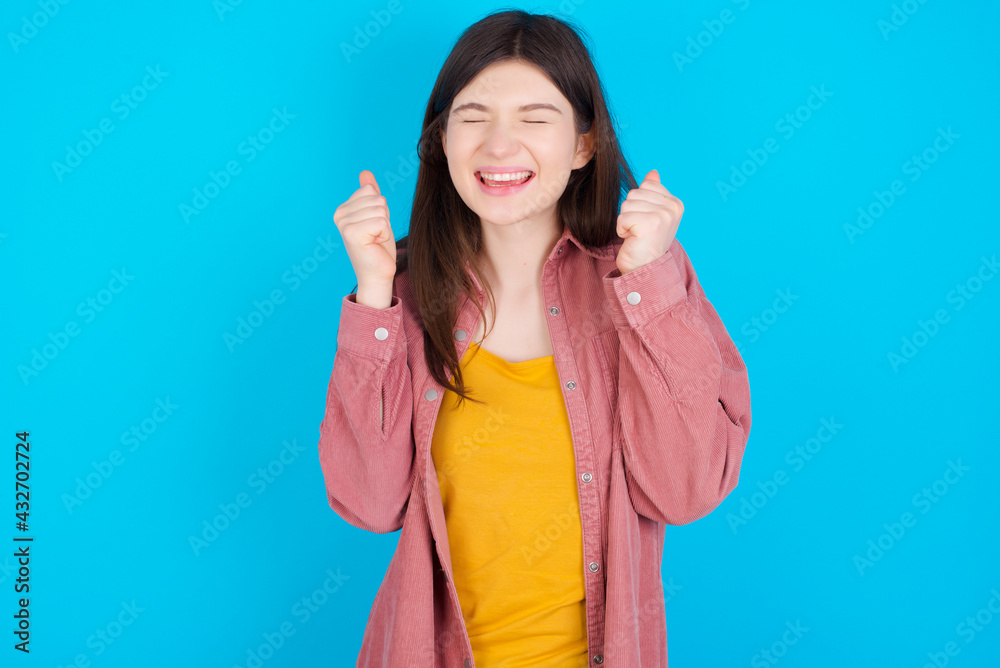 young beautiful Caucasian woman wearing pink jacket over blue wall rejoicing his success and victory clenching fists with joy being happy to achieve aim and goals. Positive emotions, feelings. young beautiful Caucasian woman wearing pink jacket over blue wall rejoicing his success and victory clenching fists with joy being happy to achieve aim and goals. Positive emotions, feelings.