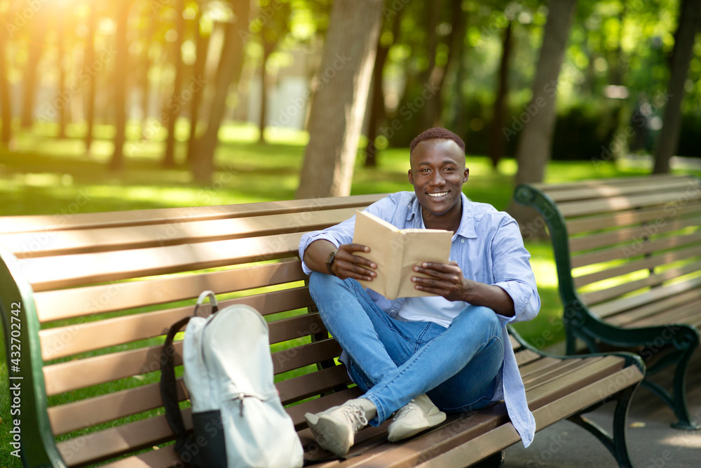 Fototapeta premium Funky African American guy reading fascinating book on bench at urban park, empty space