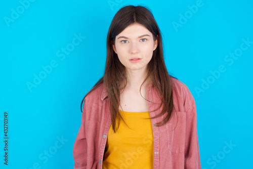 Joyful young beautiful Caucasian woman wearing pink jacket over blue wall looking to the camera, thinking about something. Both arms down, neutral facial expression.