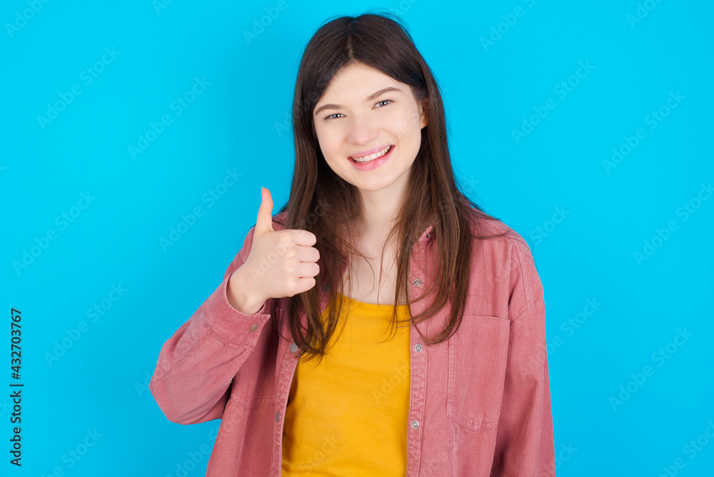 young beautiful Caucasian woman wearing pink T-shirt over blue wall giving thumb up gesture, good Job! Positive human emotion facial expression body language. young beautiful Caucasian woman wearing pink T-shirt over blue wall giving thumb up gesture, good Job! Positive human emotion facial expression body language.