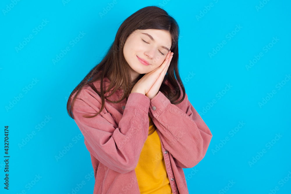 Relax and sleep time. Tired young beautiful Caucasian woman wearing pink jacket over blue wall with closed eyes leaning on palms making sleeping gesture.