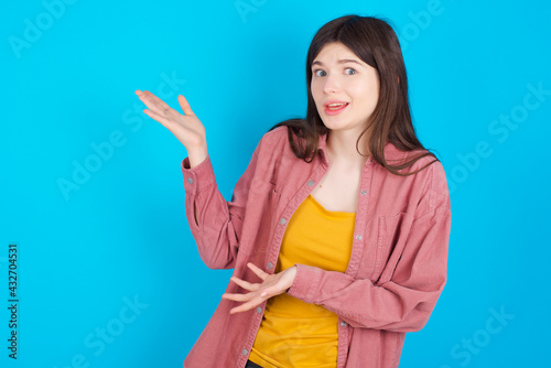 young beautiful Caucasian woman wearing pink jacket over blue wall pointing aside with both hands showing something strange and saying: I don't know what is this. Advertisement concept.