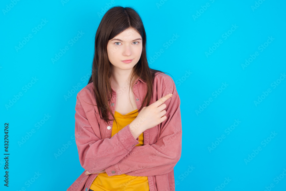 young beautiful Caucasian woman wearing pink jacket over blue wall smiling broadly at camera, pointing fingers away, showing something interesting and exciting. young beautiful Caucasian woman wearing pink jacket over blue wall smiling broadly at camera, pointing fingers away, showing something interesting and exciting.