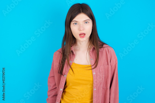 Emotional attractive young beautiful Caucasian woman wearing pink jacket over blue with opened mouth expresses great surprise and fright, stares at camera. Unexpected shocking news and human reaction.