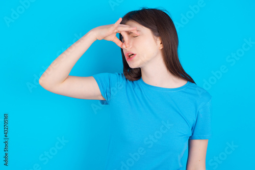 Displeased young beautiful Caucasian woman wearing blue T-shirt over blue wall plugs nose as smells something stink and unpleasant, feels aversion, hates disgusting scent.