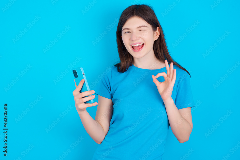 Happy young beautiful Caucasian woman wearing blue T-shirt over blue wall sending a message on his smartphone or taking a selfie  and making ok sign with his hand.
