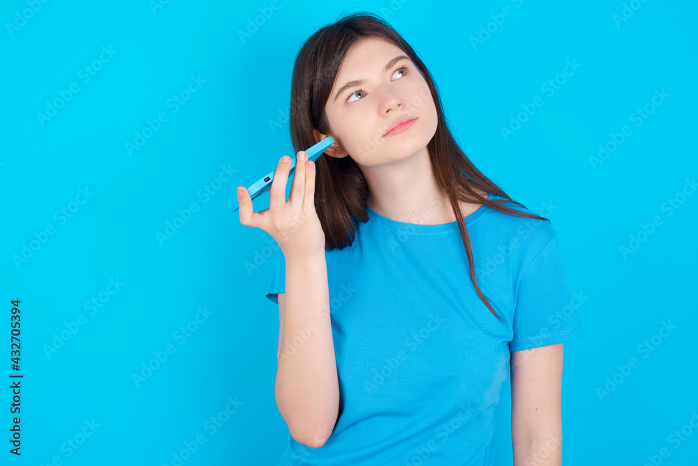 Smiling young beautiful Caucasian woman wearing blue T-shirt over blue wall listening a voice message from her smartphone. Communication and technology concept. Smiling young beautiful Caucasian woman wearing blue T-shirt over blue wall listening a voice message from her smartphone. Communication and technology concept.