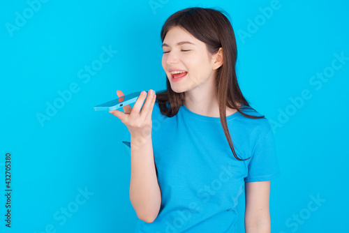 Smiling young beautiful Caucasian woman wearing blue T-shirt over blue wall sending voice message on her smart phone. Communication and new technologies concept.