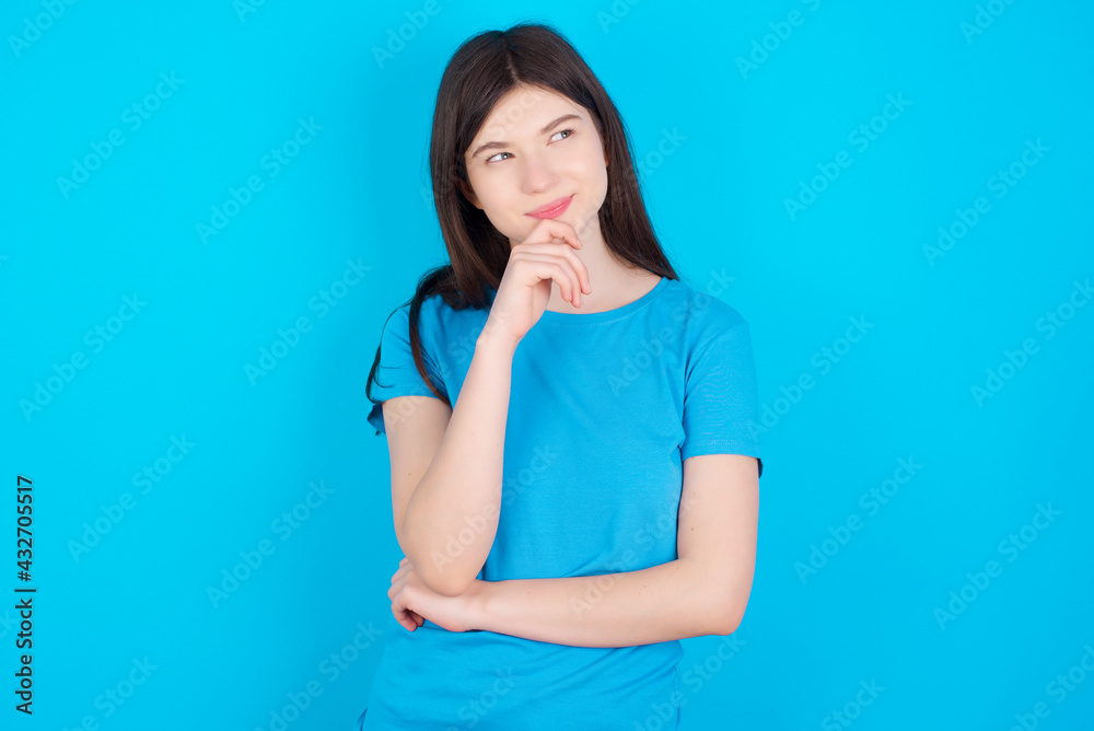 Thoughtful young beautiful Caucasian woman wearing blue T-shirt over blue wall holds chin and looks away pensively makes up great plan Thoughtful young beautiful Caucasian woman wearing blue T-shirt over blue wall holds chin and looks away pensively makes up great plan