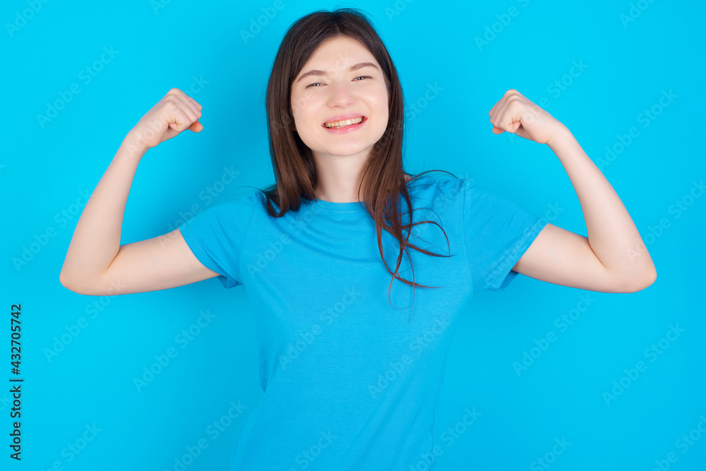 Strong powerful young beautiful Caucasian woman wearing blue T-shirt over blue wall toothy smile, raises arms and shows biceps. Look at my muscles! Strong powerful young beautiful Caucasian woman wearing blue T-shirt over blue wall toothy smile, raises arms and shows biceps. Look at my muscles!